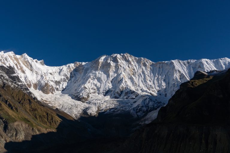 Snow-covered mountain peaks under a clear blue sky, casting deep shadows on the rugged slopes below.