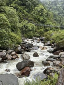 Rocky river with white rapids, lush green banks, and a suspension bridge spanning the valley in the distance.
