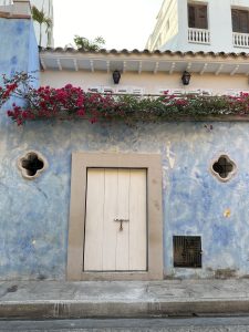 Old white door on a weathered blue wall with a decorative window and pink bougainvillea flowers.