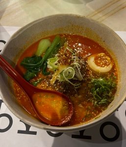 Bowl of spicy noodle soup with soft-boiled egg, green onions, sesame seeds, and leafy greens.