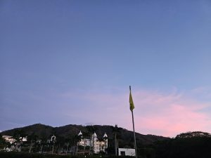 A white resort-style building with palm trees stands at the base of a forested hill beneath a pink and blue evening sky. A tall flagpole rises in the foreground