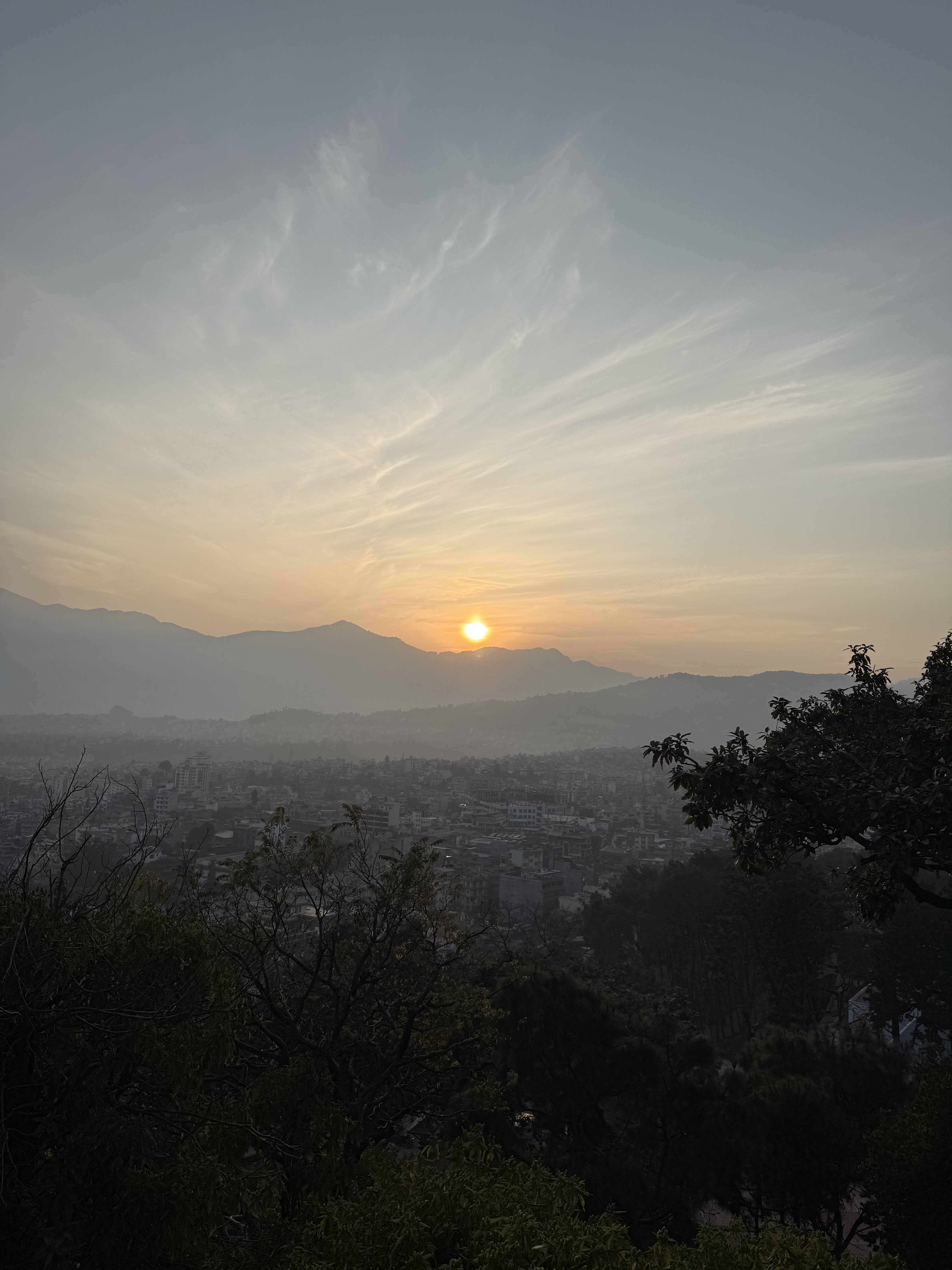 Sunset over a cityscape with distant mountains and wispy clouds.