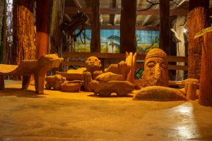 A collection of carved stone sculptures depicting animals and a large human head, displayed on a sandy floor inside a warmly lit wooden structure with a painted mural of tropical birds in the background.
