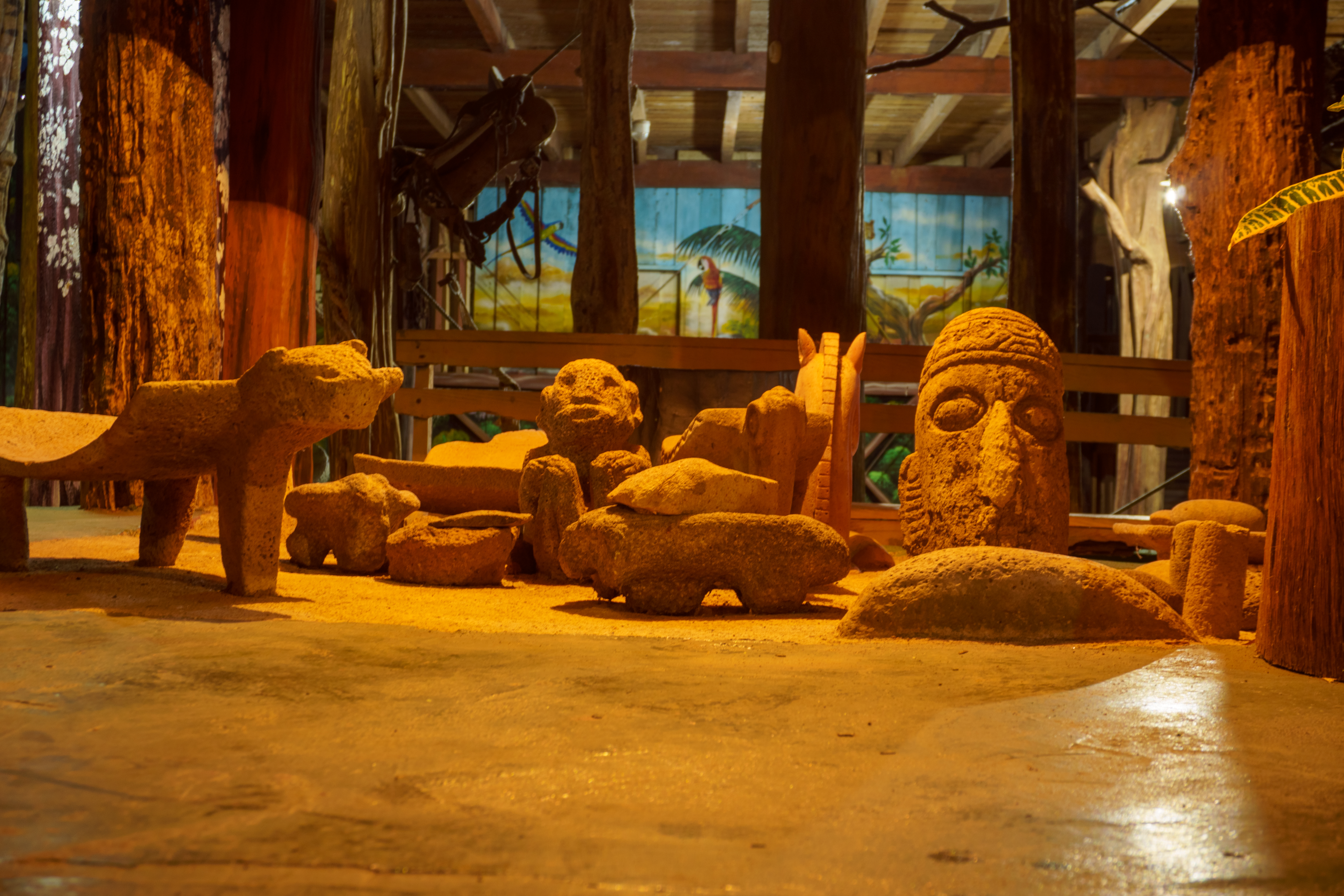 A collection of carved stone sculptures depicting animals and a large human head, displayed on a sandy floor inside a warmly lit wooden structure with a painted mural of tropical birds in the background.