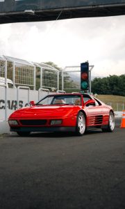 Low-angle shot of a bright red Ferrari 348 Spider with a tan leather interior, positioned on an asphalt racetrack next to a drag racing starting light and a safety fence , all at a Wekfest event in Autódromo de Costa Rica. 