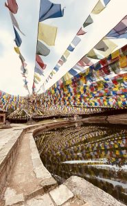 A peaceful temple courtyard adorned with vibrant Buddhist prayer flags, beautifully reflected in a calm stone pond beneath a bright sky.