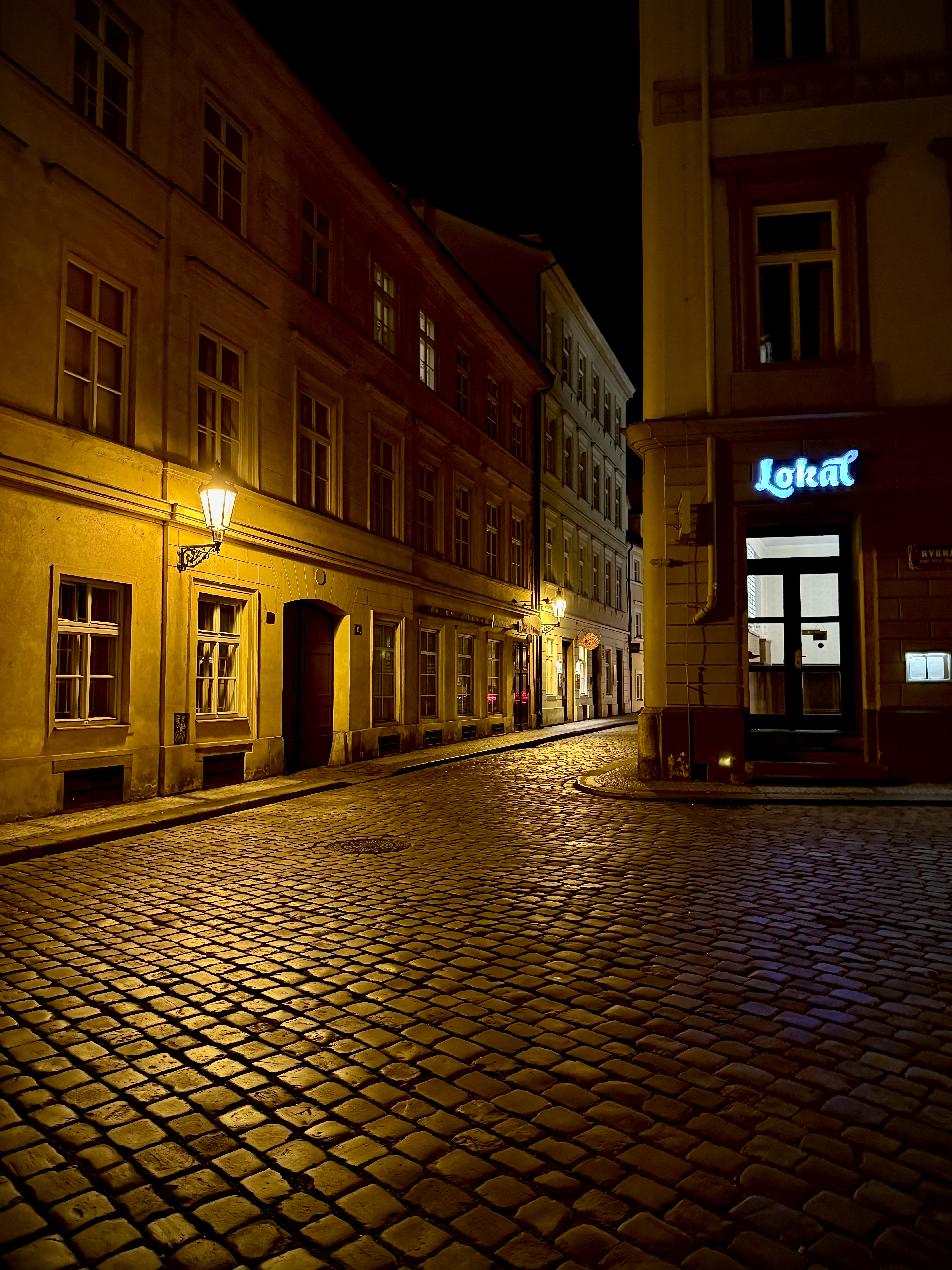 Cobbled street illuminated by historic street lamps between old buildings at night in Prague, Czech Republic.