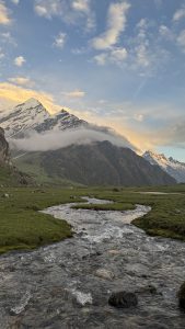 A serene landscape featuring a clear stream flowing through a green meadow with snow capped mountains in the background under a sky with scattered clouds.