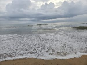 Sea foam covers wet sand as gentle waves roll onto the shore under a cloudy sky.
