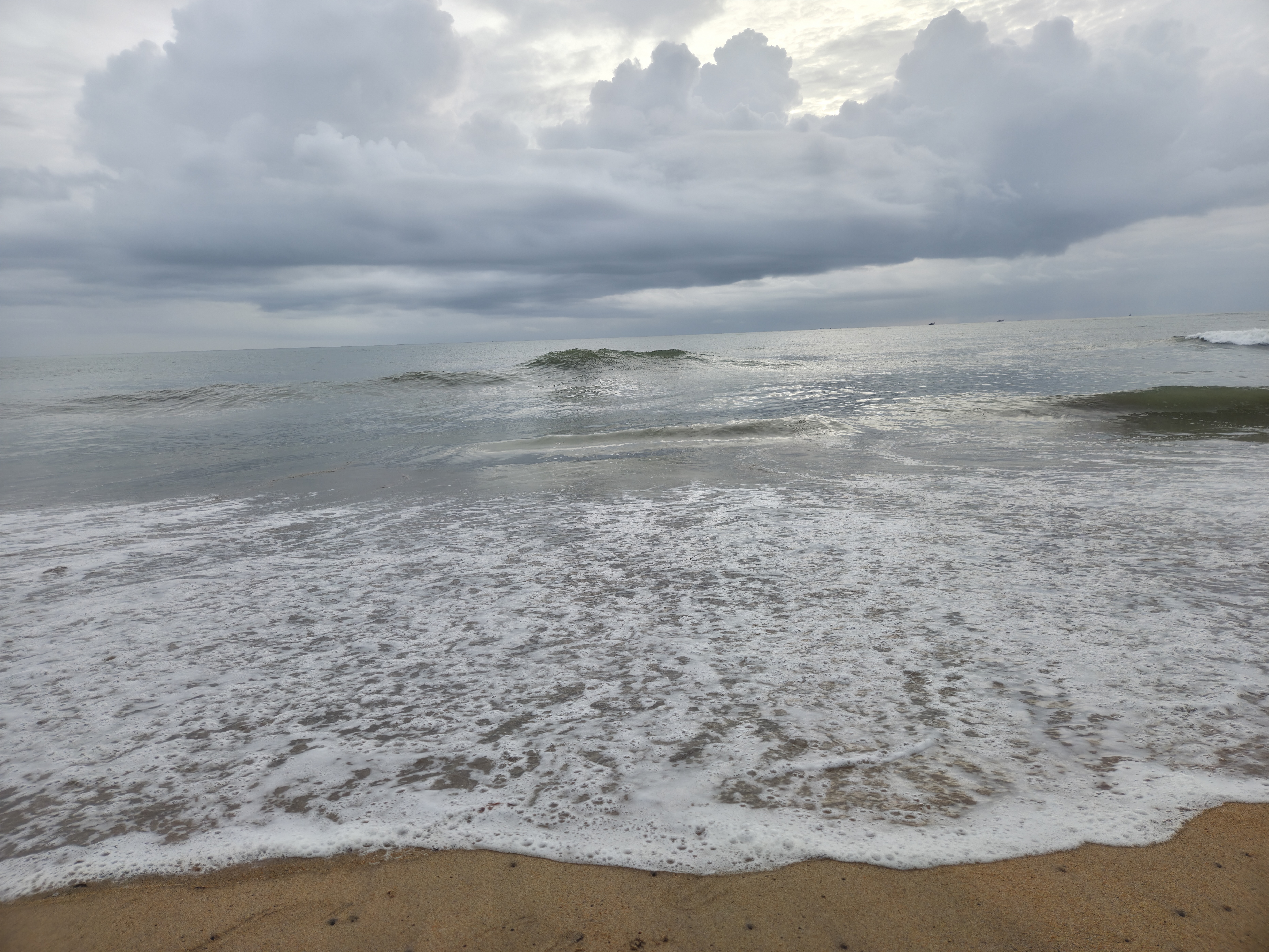Sea foam covers wet sand as gentle waves roll onto the shore under a cloudy sky.