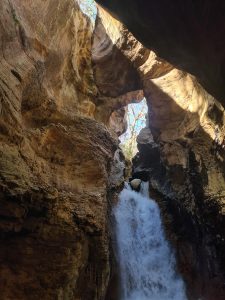 A waterfall flows between tall, rocky canyon walls. Sunlight enters through openings above the rocks and illuminates parts of the cave-like space. The scene shows water, stone textures, and natural erosion.