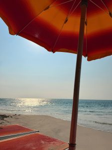 A striped orange and red beach umbrella on a sandy beach with the ocean in the background.