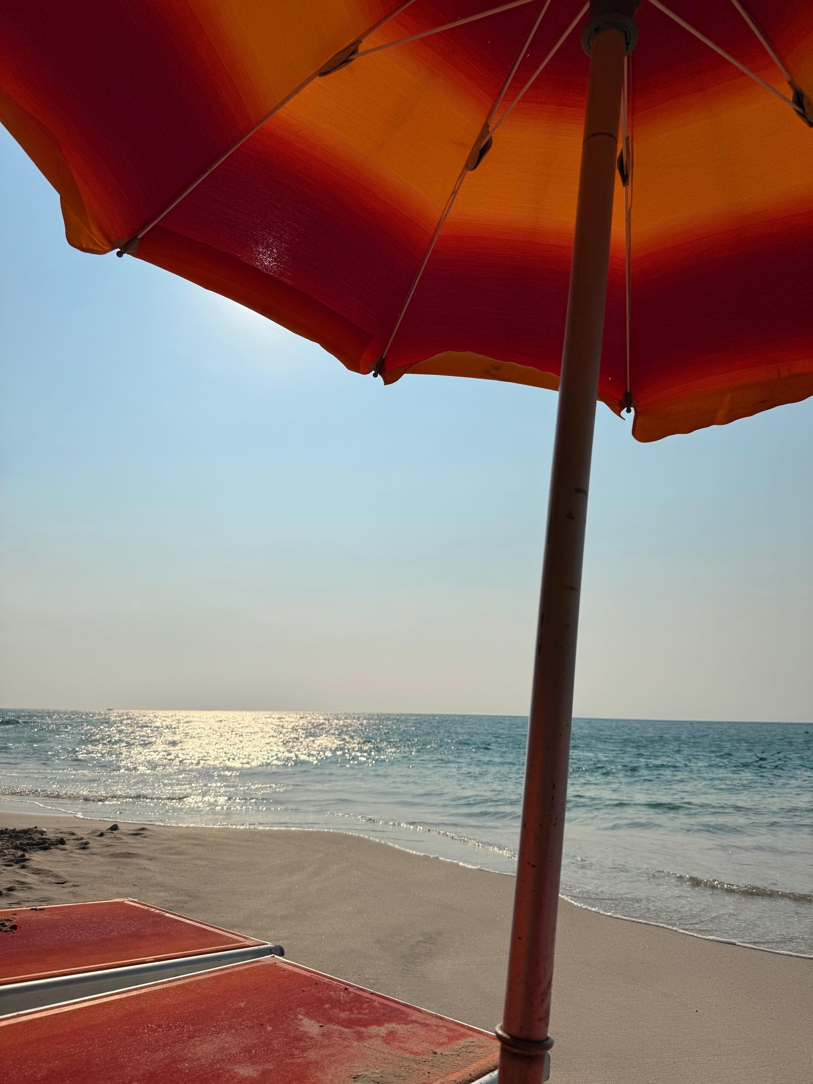 A striped orange and red beach umbrella on a sandy beach with the ocean in the background.