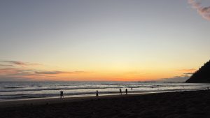 
Sunset beach with gentle waves, walking silhouettes, and hills in the background.