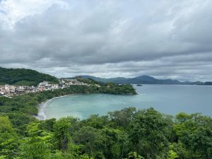Panoramic view of a crescent-shaped beach, a clear blue-green bay, and white buildings among green hills.