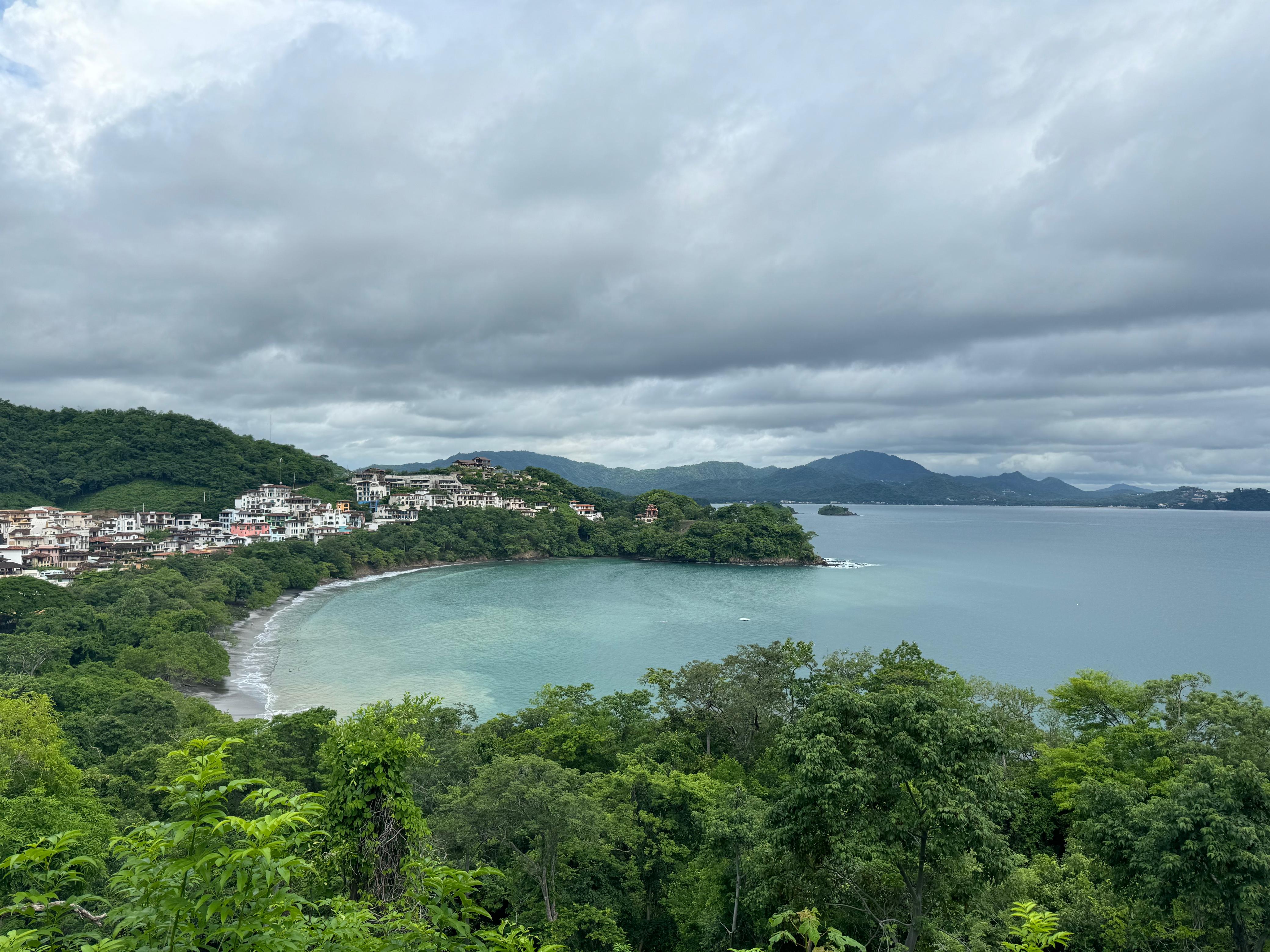 Panoramic view of a crescent-shaped beach, a clear blue-green bay, and white buildings among green hills.