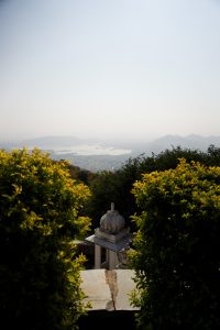 A scenic view shows a domed pavilion between lush green bushes, overlooking a vast landscape with lakes and distant mountains under a clear sky.