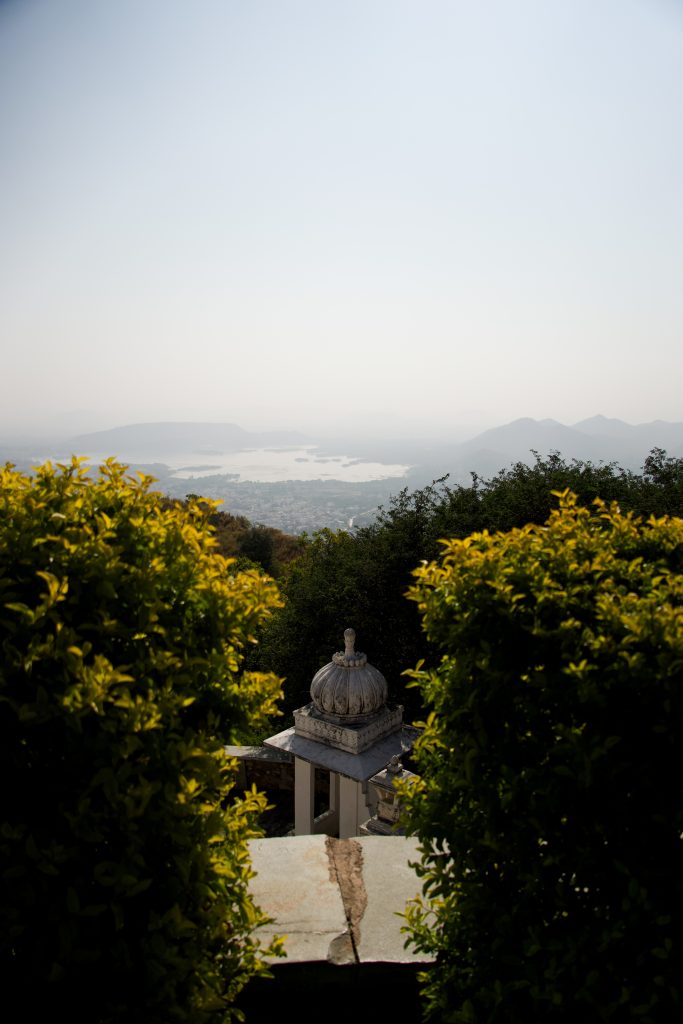 A scenic view shows a domed pavilion between lush green bushes, overlooking a vast landscape with lakes and distant mountains under a clear sky.