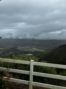 
Panoramic view of green mountains with a winding river, a white fence, and cloudy skies.