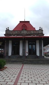 Front view of the historic Ferro-Carril al Atlántico train station in Costa Rica, showing its red pyramidal roof, decorative wooden balustrade and covered platform entrance.