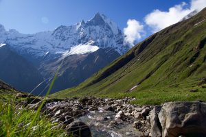 Rocky stream in a green valley with a jagged snow-capped peak rising above grassy slopes