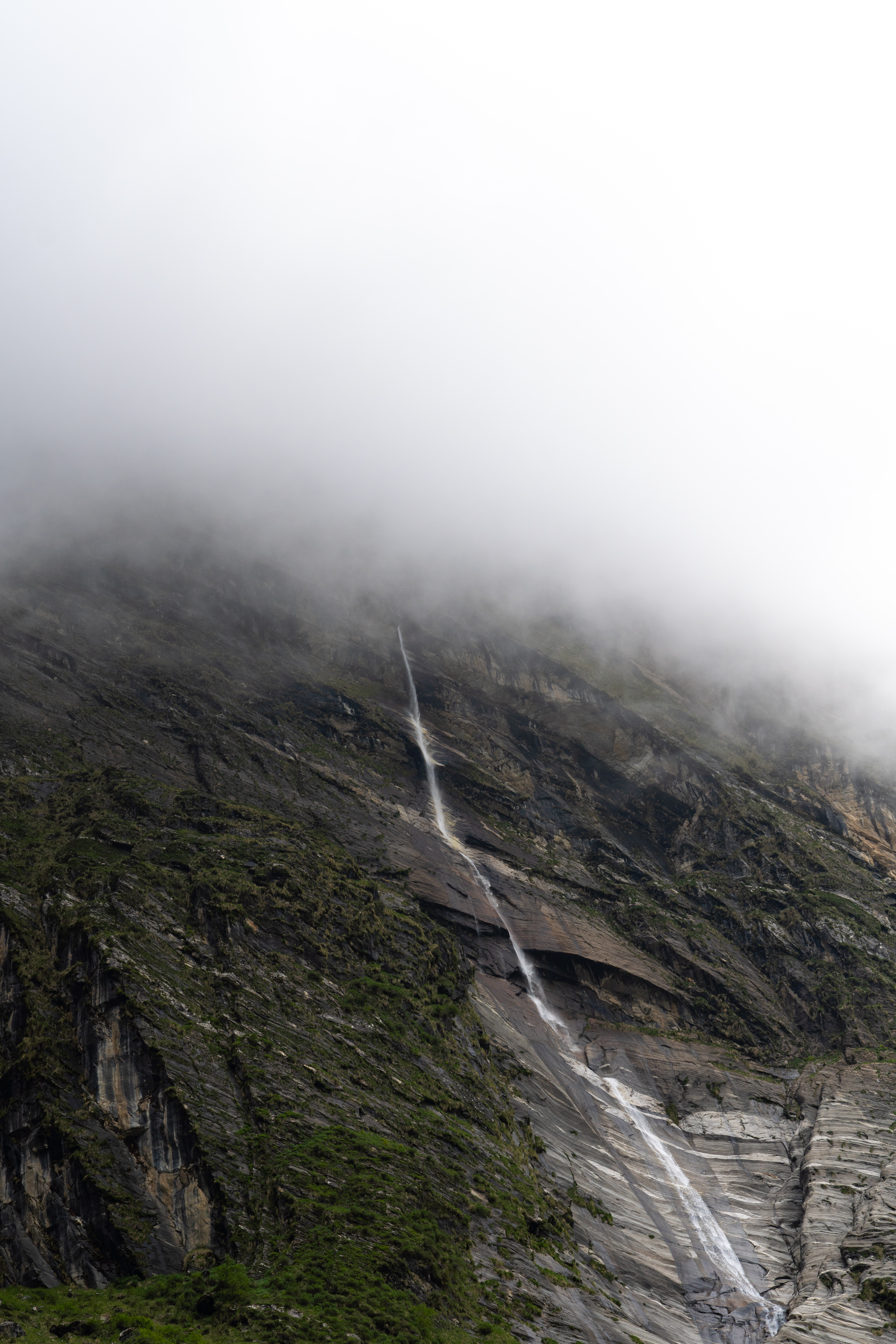 Thin waterfall streaming down a steep rocky mountain slope, upper section obscured by fog