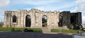 The historic ruins of the Santiago Apostle Church rise against a partly cloudy sky in Cartago, Costa Rica. The ancient stone walls, darkened with age, feature large arched openings that once served as windows and doorways. The ruins stand as a powerful reminder of the earthquakes that have shaped the history of the region.