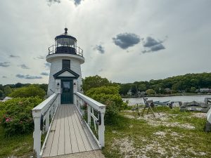A two-story lighthouse with a long ramp up to it, and the sun’s rays in the sky and clouds behind it.
