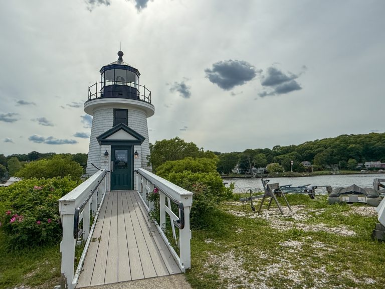 A two-story lighthouse with a long ramp up to it, and the sun’s rays in the sky and clouds behind it.