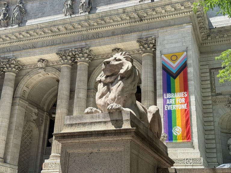 One of the stone lions outside the New York Public Library, with a perpendicular pride banner that says “Libraries are for Everyone” with the lion logo at the bottom of it.