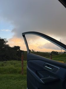 View from an open car door in Coronado, overlooking green fields and trees beneath dramatic gray clouds illuminated by soft golden sunset light.
