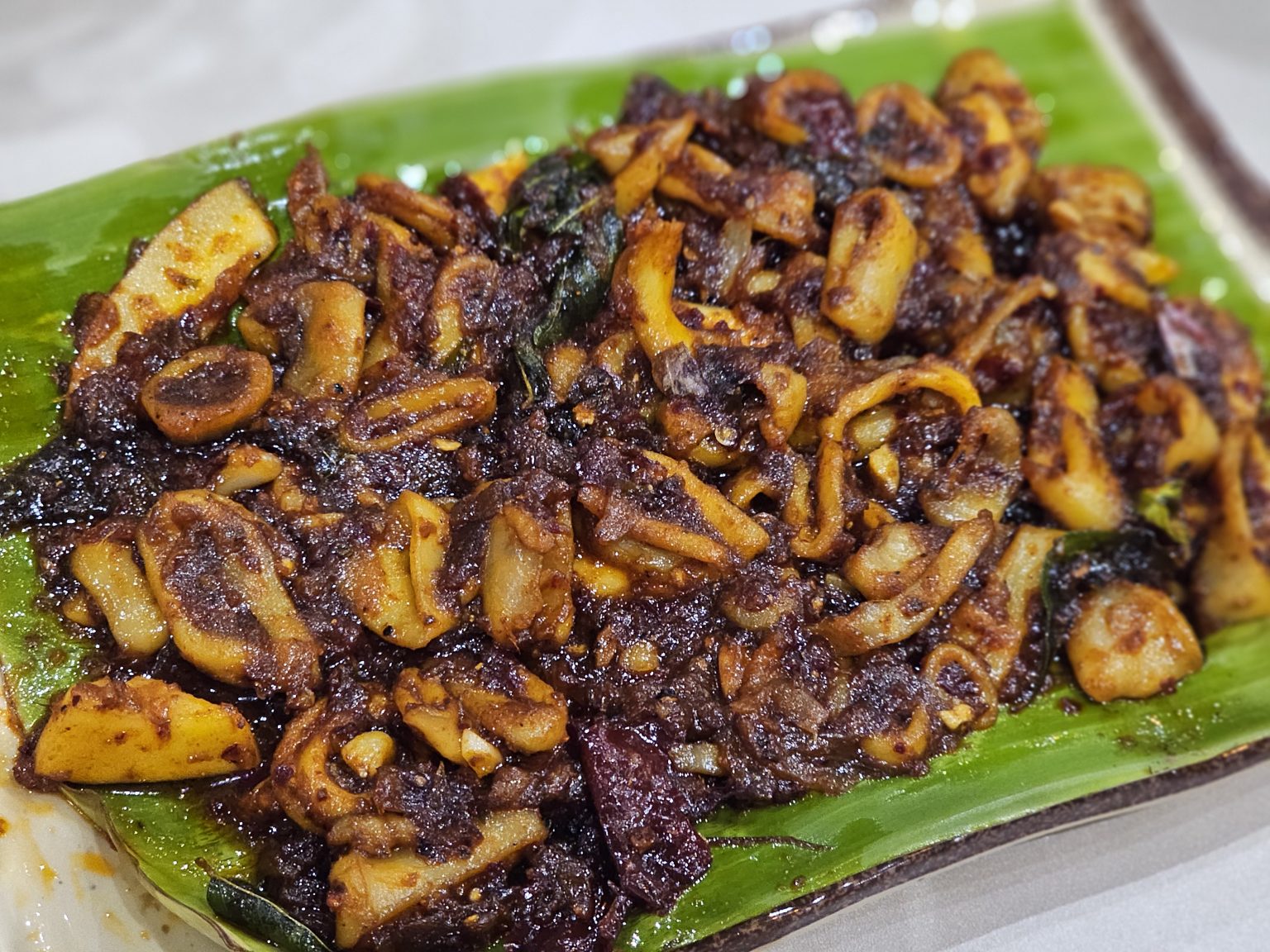A flavorful Kerala-style squid roast served on a fresh banana leaf, cooked with spices, onions, curry leaves, and roasted coconut pieces. Rich in color and texture. Captured in Nadakkavu, Kozhikode.