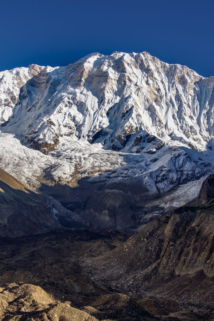Massive snow-covered mountain wall above a rocky moraine valley under a clear dark blue sky