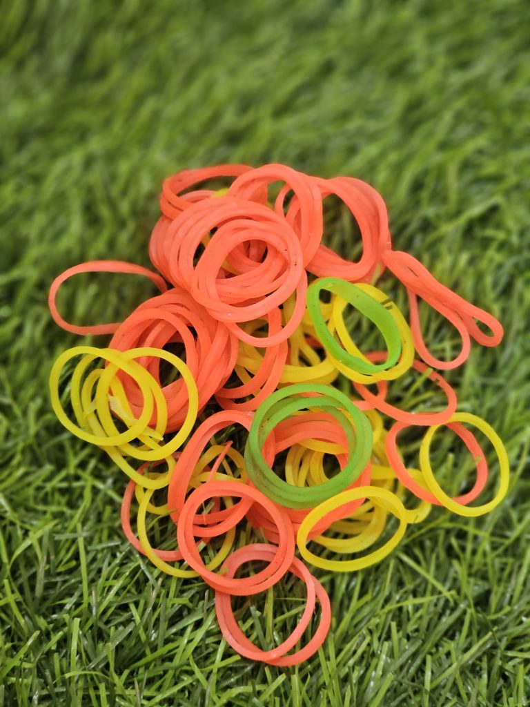 A pile of colorful rubber bands rests on fresh green grass, creating a bright contrast of colors and simple shapes. The close-up view highlights the playful arrangement and vibrant tones. Captured in Perumanna, Kozhikode.