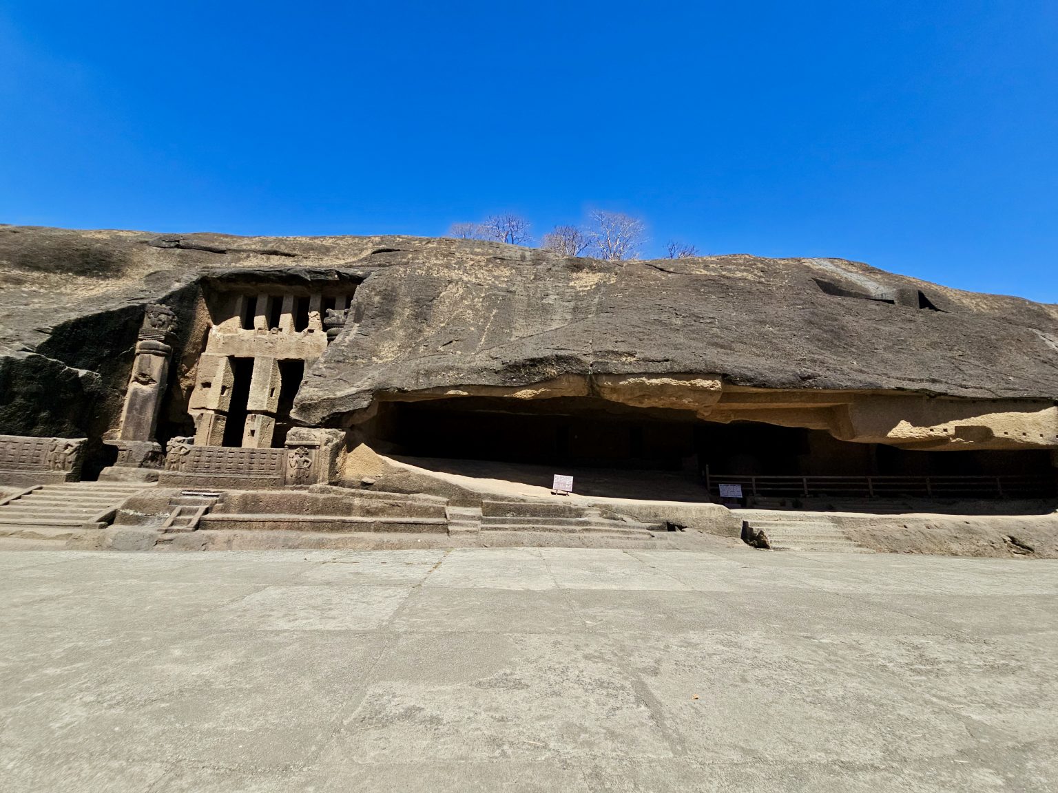 A wide view of the ancient Kanheri Caves carved into a massive basalt hill. The open courtyard, rock-cut architecture, and bright blue sky highlight the beauty of this historic Buddhist site. Captured at Kanheri Caves, Borivali East, Mumbai.