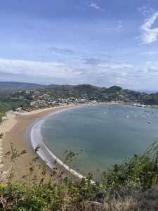Panoramic view of a curved beach with golden sand, turquoise water, anchored boats, green hills, and a small coastal town in the distance beneath a partly cloudy sky.