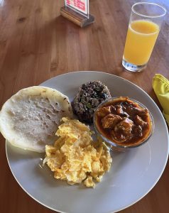 A white plate with scrambled eggs, rice and black beans, a tortilla, and a bowl of meat stew.