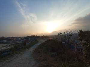 A winding dirt path climbs a grassy hillside overlooking a hazy city at sunset in kathmandu, with golden light, wispy clouds, and a peaceful evening sky.