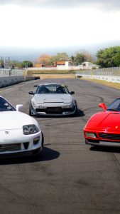 Low-angle shot of a car lineup on the main straight of Autódromo de Costa Rica. A gray Nissan Silvia drift car with pop-up headlights sits centered in the background. In the foreground, the front ends of a white Toyota Supra on the left and a bright red Ferrari 348 GTS on the right frame the shot on the asphalt track under a bright sky.