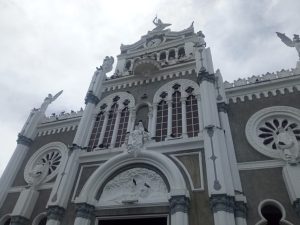 Facade of the Basilica of Our Lady of the Angels in Cartago, Costa Rica. Byzantine-Romanesque style with grey stone and white trim, a central clock tower, rose windows, arched openings and angel statues crowning the roofline.