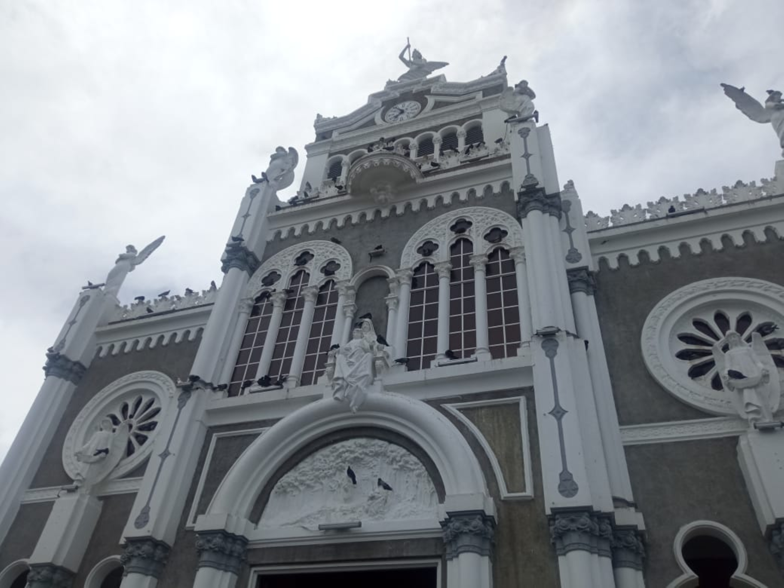 Facade of the Basilica of Our Lady of the Angels in Cartago, Costa Rica. Byzantine-Romanesque style with grey stone and white trim, a central clock tower, rose windows, arched openings and angel statues crowning the roofline.