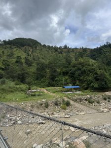 A view through the wire mesh of a suspension bridge shows a muddy river flowing below. On the grassy bank beyond, a dirt path winds past two simple shelters—one with a bright blue roof. In the background, a steep, lush green hill covered in dense forest rises toward a cloudy sky.
