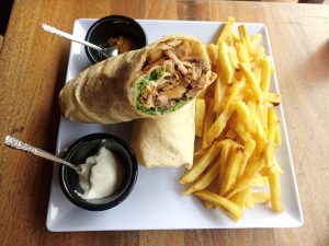 A mixed beef and chicken shawarma cut in half and served with French fries, accompanied by two dipping sauces on a white square plate.
