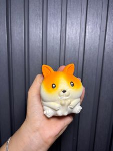 A hand holding a small orange and white fox-shaped sensory squeeze toy with a rounded design, photographed against a dark gray corrugated metal wall with even lighting.