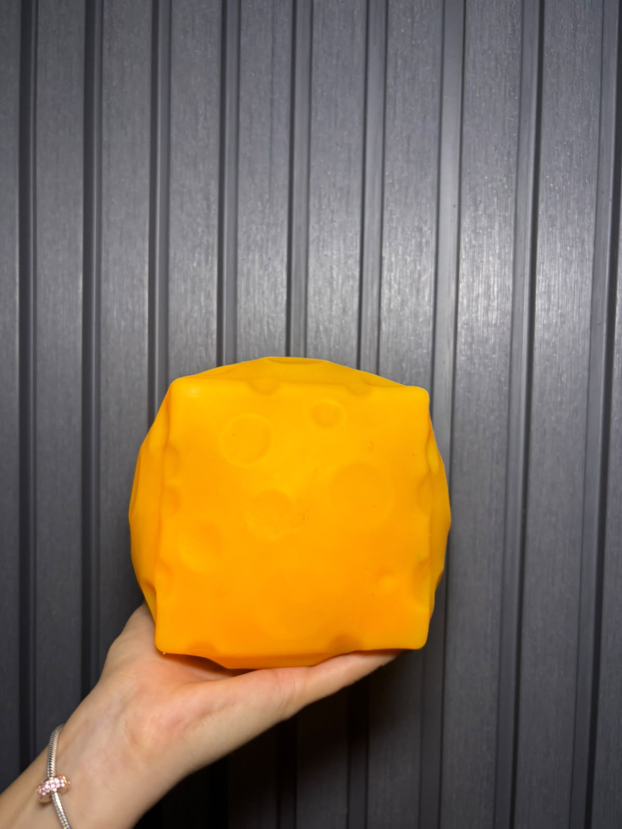 A hand holding an orange textured sensory cube with a soft geometric design, photographed against a dark gray corrugated metal background with even lighting.