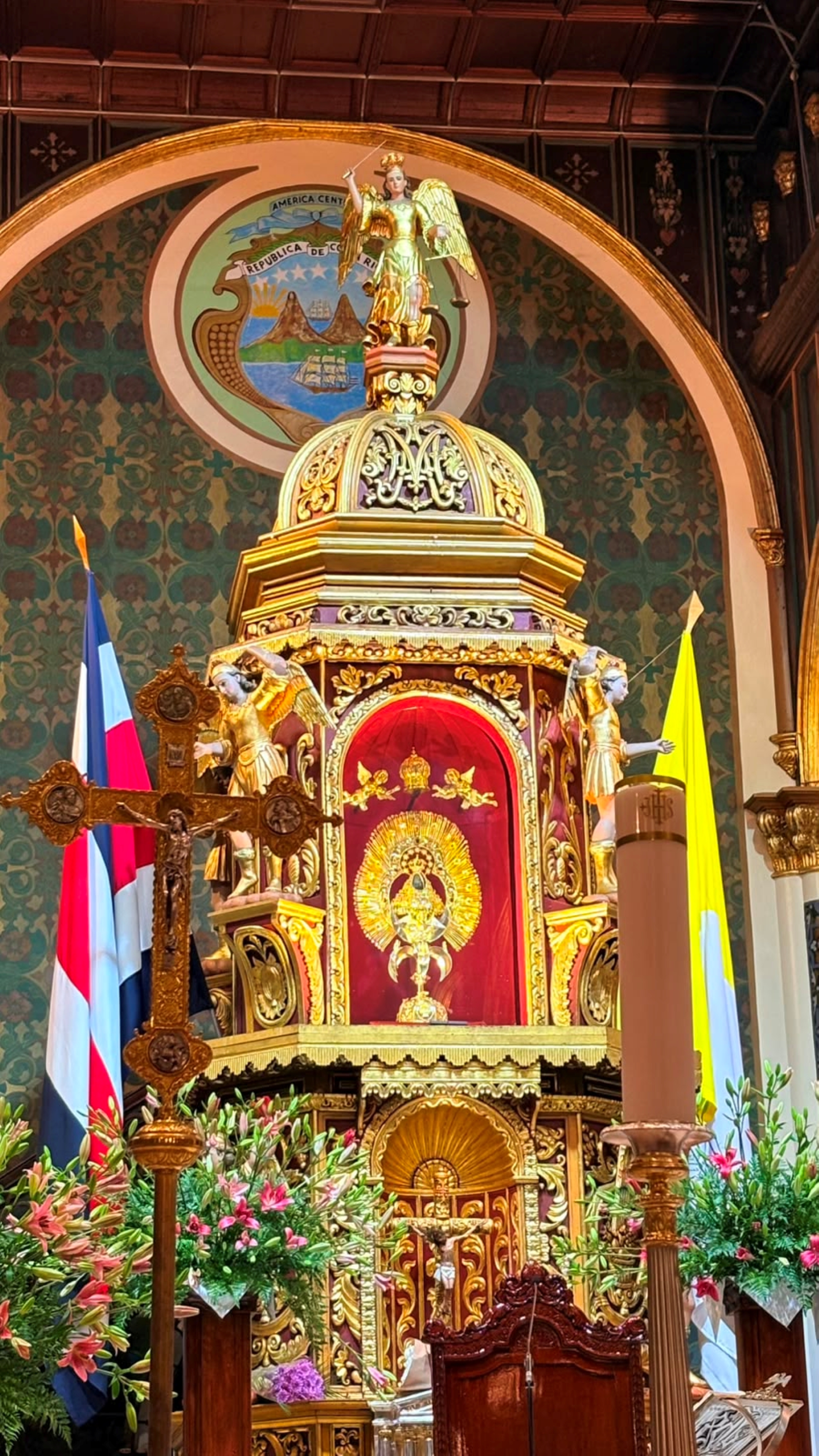 Golden ornate altar housing La Negrita, the Virgin of Los Angeles, at the Basilica in Cartago, Costa Rica. Gilded angel figures, the national coat of arms and a crucifix surround the sacred image, framed by lilies and the Costa Rican flag.
