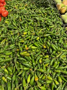 A large pile of fresh green chilly peppers at a market, with red tomatoes on the left and light green pears on the right.