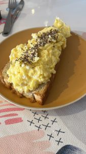 A slice of toasted bread topped with scrambled eggs sits on a round yellow plate. The eggs are soft and fluffy, with a bright yellow color, and are sprinkled with a mix of seeds, including black sesame seeds and flaxseeds, adding visual contrast. 