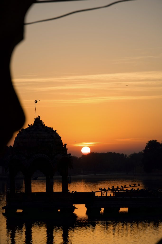 Sunset view over a tranquil lake with an ornate gazebo silhouette in the foreground. The sky is vibrant, with hues of orange and gold reflecting on the water.