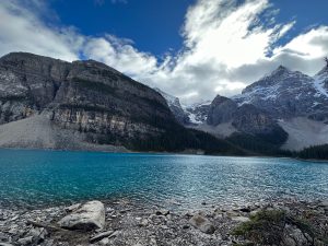 Turquoise water of Moraine Lake stretches beneath towering rocky mountains dusted with snow, while dark evergreen forest lines the shore. A rocky foreground leads to the rippling lake, and dramatic white clouds drift across a vivid blue sky, creating a striking Canadian Rockies landscape scene.
